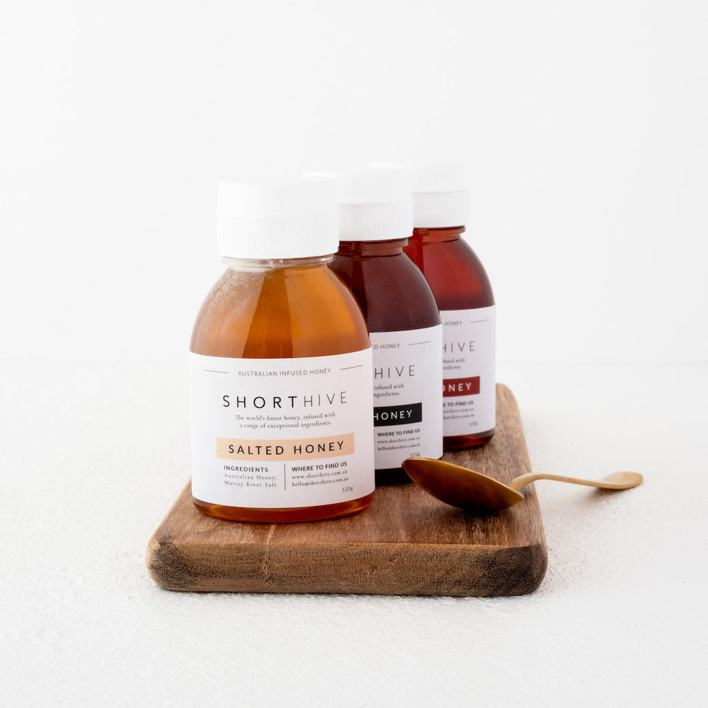 125g of bottles of Salted (caramel) honey, Espresso (coffee) honey, Chilli (spicy) honey pictured on a wooden chopping board with gold spoon placed beside thee board.