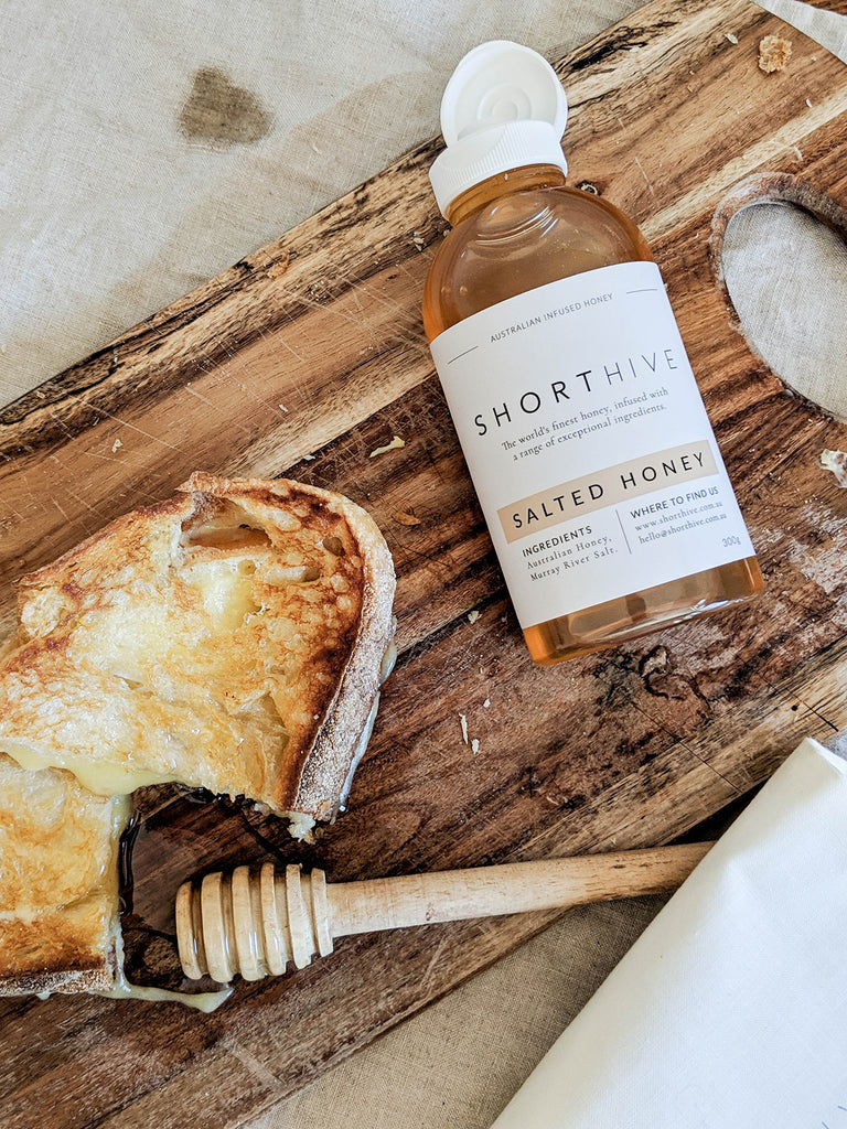ShortHive Australian Caramel Salted Honey bottle open pictured on a wooden cutting board with a grilled cheese oozing with cheese and salted honey.