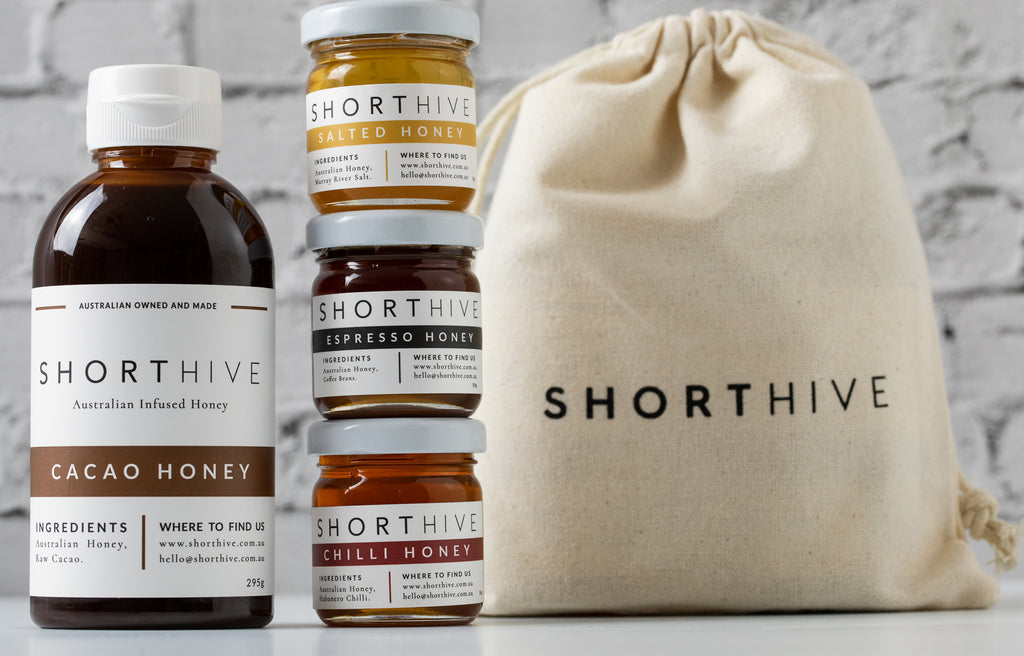 ShortHive Australian Cacao (chocolate) Infused Honey pictured with calico bag and three stacked mini 50g bottles of Espresso (coffee) honey, Salted (caramel) honey, Chilli (spicy) honey. 
